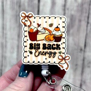 May include: A fall-themed badge reel with the text "Big Back Energy." The design features illustrations of a pumpkin spice latte, pumpkin pie, a roasted turkey, and a cupcake topped with a pumpkin. The badge reel has a white retractable cord and a clear plastic clip.