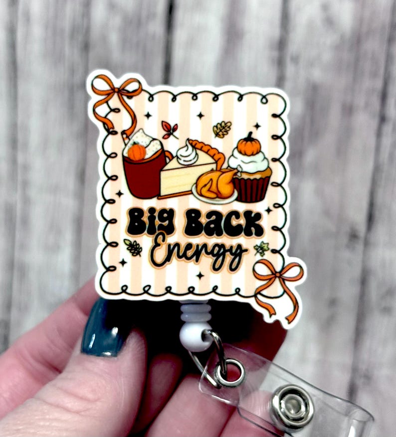 May include: A fall-themed badge reel with the text "Big Back Energy." The design features illustrations of a pumpkin spice latte, pumpkin pie, a roasted turkey, and a cupcake topped with a pumpkin. The badge reel has a white retractable cord and a clear plastic clip.