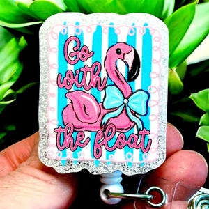 May include: A badge reel with a pink flamingo design. The flamingo has a blue bow and is set against a blue and white striped background. The text "Go with the float" is written in pink script.