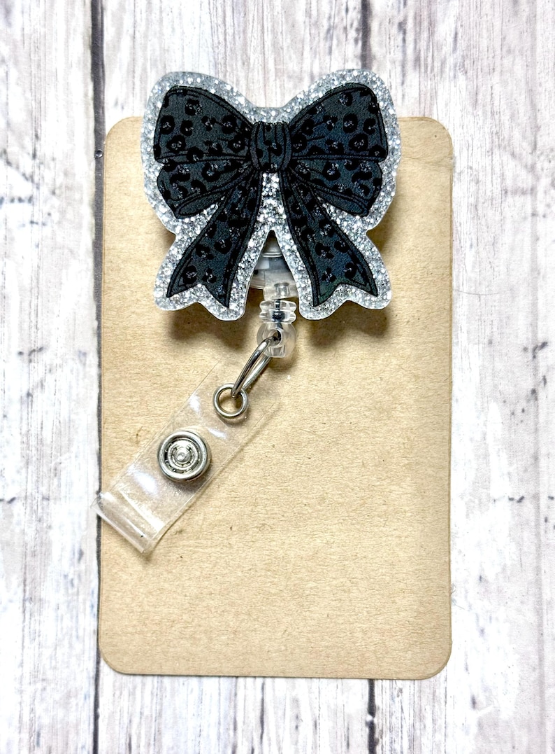 Puede incluir: Un carrete de insignia en forma de lazo negro con un contorno de purpurina plateada y un dise&ntilde;o de estampado de leopardo negro. El carrete de insignia tiene una correa de pl&aacute;stico transparente y un clip met&aacute;lico. El carrete de insignia est&aacute; sujeto a una tarjeta rectangular de color canela.