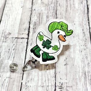 May include: A white duck wearing a green hat and holding a shamrock. The duck is wearing green boots and is attached to a clear plastic badge holder.
