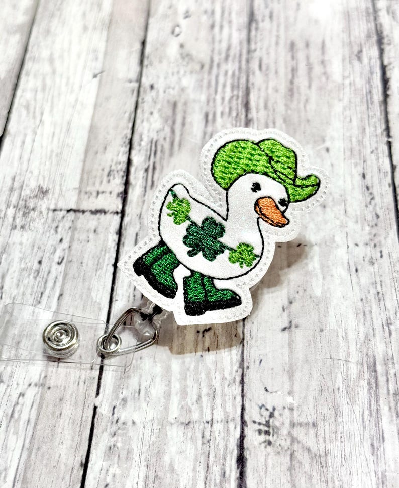 May include: A white duck wearing a green hat and holding a shamrock. The duck is wearing green boots and is attached to a clear plastic badge holder.
