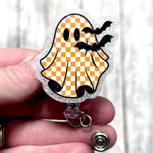 May include: A Halloween-themed badge reel featuring a ghost with an orange and white checkered pattern. The ghost has black eyes and is accompanied by three black bats. The badge reel has a clear, glittery border and a retractable cord.
