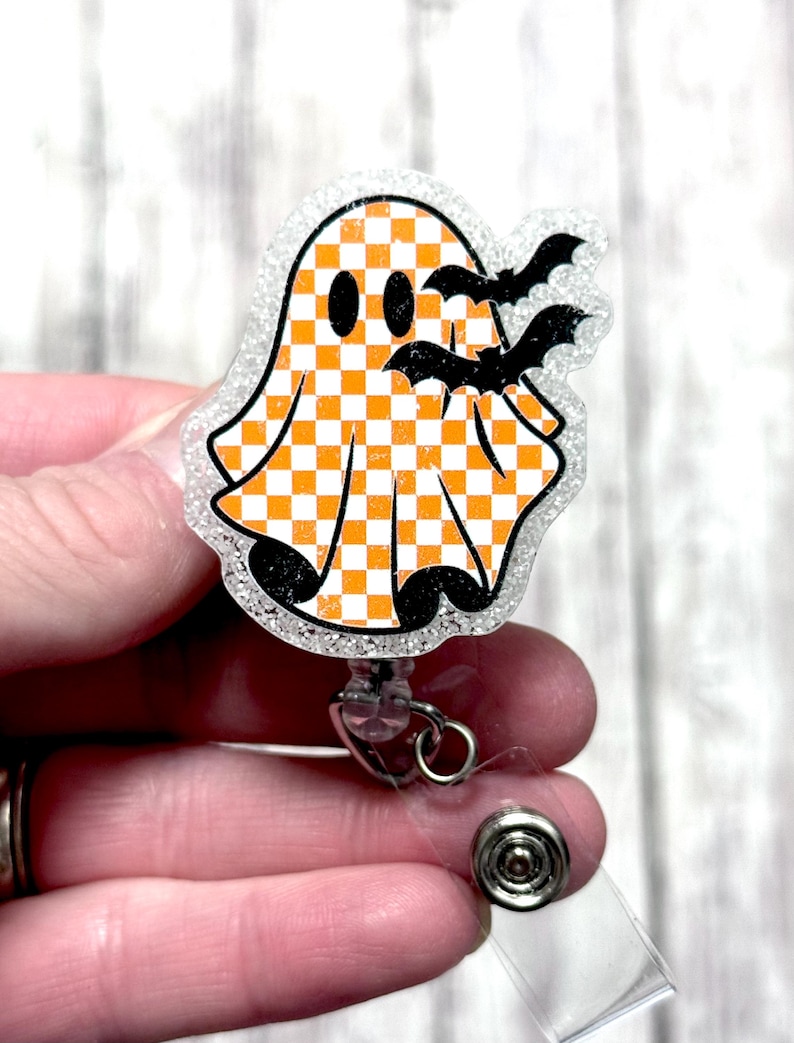 May include: A Halloween-themed badge reel featuring a ghost with an orange and white checkered pattern. The ghost has black eyes and is accompanied by three black bats. The badge reel has a clear, glittery border and a retractable cord.