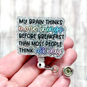 May include: A glittery badge reel with the quote, "MY BRAIN THINKS more things BEFORE BREAKFAST THAN MOST PEOPLE THINK all day." The text is in black and iridescent colours. The reel has a white plastic base, a metal clip, and a clear plastic strap.
