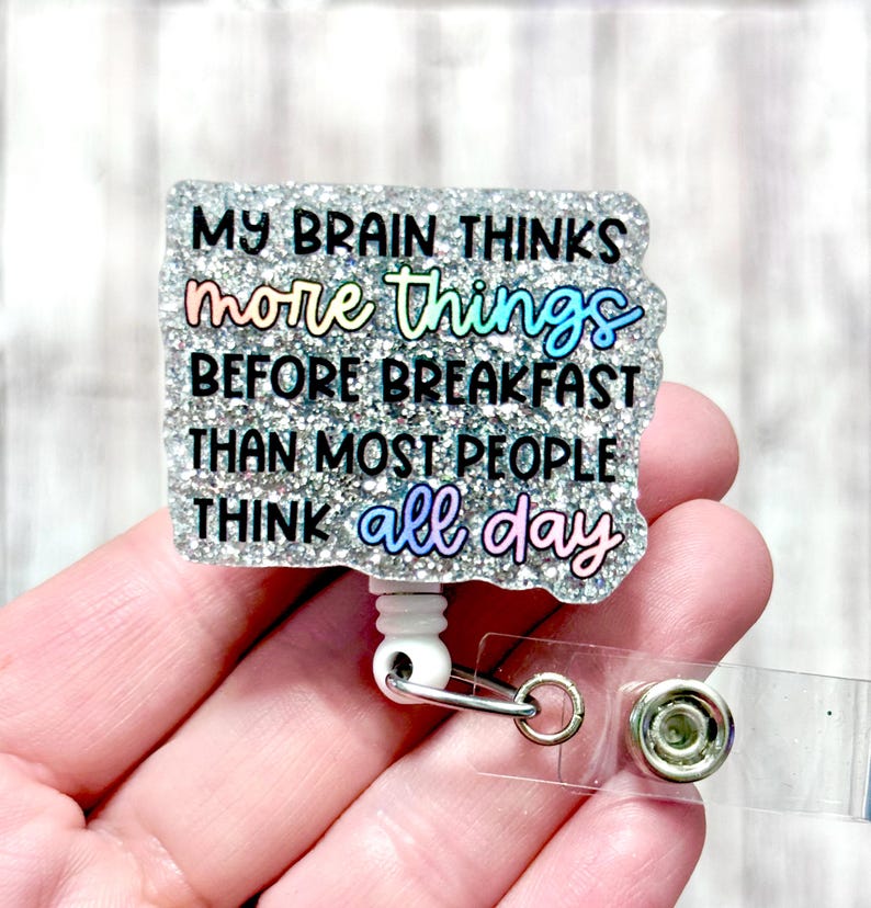 May include: A glittery badge reel with the quote, "MY BRAIN THINKS more things BEFORE BREAKFAST THAN MOST PEOPLE THINK all day." The text is in black and iridescent colours. The reel has a white plastic base, a metal clip, and a clear plastic strap.