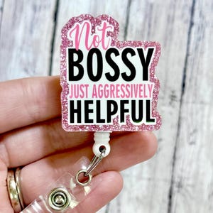 May include: A pink glitter badge reel with the text "Not Bossy Just Aggressively Helpful" in black and pink. The badge reel has a clear plastic clip and a white retractable cord.