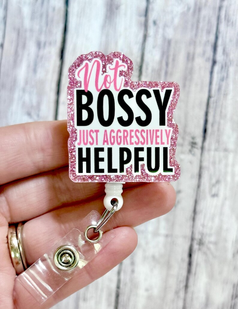 May include: A pink glitter badge reel with the text "Not Bossy Just Aggressively Helpful" in black and pink. The badge reel has a clear plastic clip and a white retractable cord.