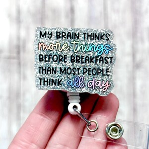 May include: A badge reel with a glittery, textured surface and the text "MY BRAIN THINKS more things BEFORE BREAKFAST THAN MOST PEOPLE THINK all day". The text is in colourful lettering. It has a white cord and a clear clip.