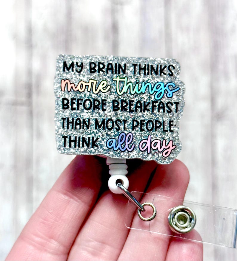 May include: A badge reel with a glittery, textured surface and the text "MY BRAIN THINKS more things BEFORE BREAKFAST THAN MOST PEOPLE THINK all day". The text is in colourful lettering. It has a white cord and a clear clip.