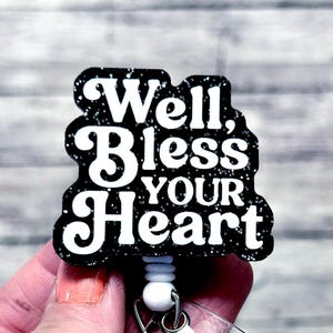 May include: A black and silver glitter badge reel with the phrase "Well, Bless Your Heart" in white lettering. The badge reel has a white retractable cord and a white plastic clip.