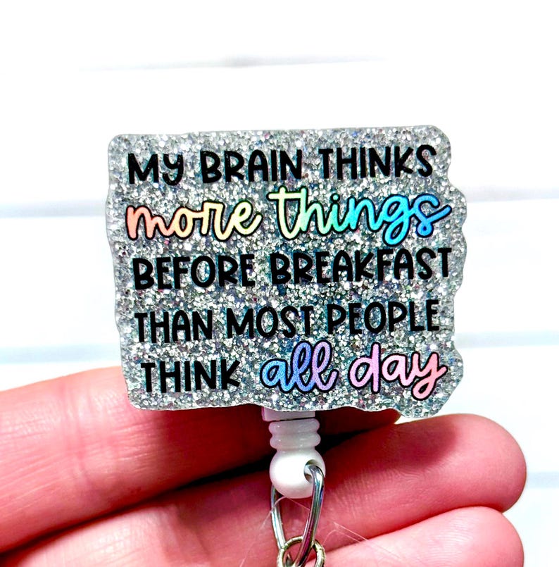 May include: A glittery badge reel with the text "MY BRAIN THINKS more things BEFORE BREAKFAST THAN MOST PEOPLE THINK all day" in black and iridescent lettering. The reel has a white plastic clip and a retractable cord.