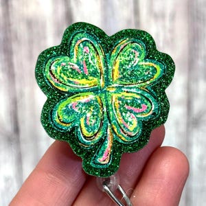 May include: A green glitter shamrock badge reel with a four-leaf clover design. The clover has yellow and pink accents. The badge reel has a silver clip and a clear plastic strap.
