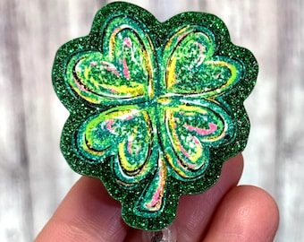 May include: A green glitter shamrock badge reel with a four-leaf clover design. The clover has yellow and pink accents. The badge reel has a silver clip and a clear plastic strap.
