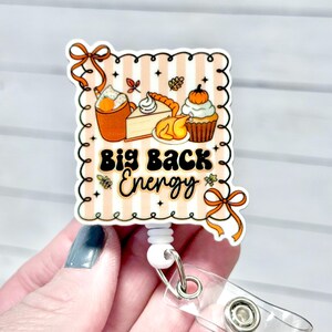 May include: A retractable badge reel with a fall-themed design. The design features a pumpkin spice latte, pie, and a cupcake with the words "Big Back Energy". The reel has a clear plastic strap and a metal snap.