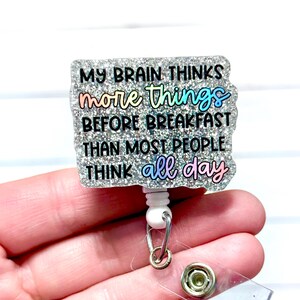 May include: A glittery badge reel with the text "MY BRAIN THINKS more things BEFORE BREAKFAST THAN MOST PEOPLE THINK all day." The text is in black and rainbow colours. The reel has a white base and a silver retractable cord.