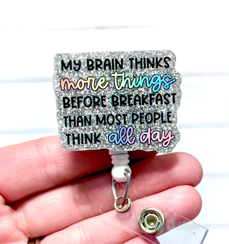 May include: A glittery badge reel with the text "MY BRAIN THINKS more things BEFORE BREAKFAST THAN MOST PEOPLE THINK all day." The text is in black and rainbow colours. The reel has a white base and a silver retractable cord.