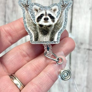 May include: A silver glitter badge reel with a raccoon image. The raccoon is standing on its hind legs with its arms raised in the air. The badge reel has a clear plastic strap and a metal clip.