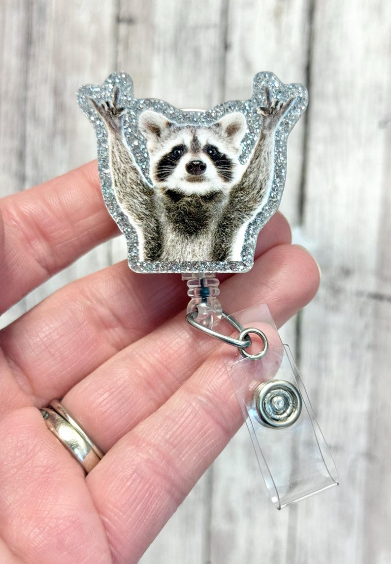 May include: A silver glitter badge reel with a raccoon image. The raccoon is standing on its hind legs with its arms raised in the air. The badge reel has a clear plastic strap and a metal clip.