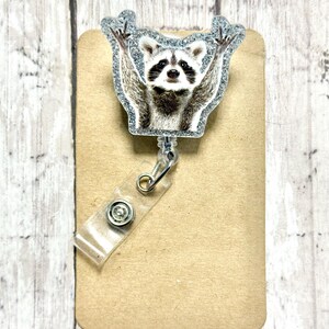 May include: A silver glitter badge reel with a raccoon image. The raccoon is holding its paws up in the air.