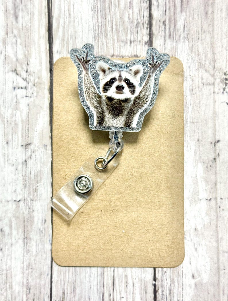 May include: A silver glitter badge reel with a raccoon image. The raccoon is holding its paws up in the air.