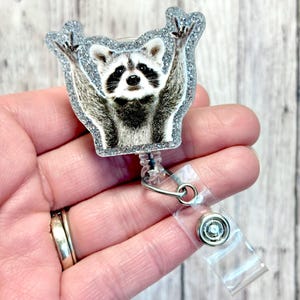 May include: A silver glitter badge reel with a raccoon wearing a surprised expression and raising its paws in the air. The raccoon is printed on a clear plastic circle.