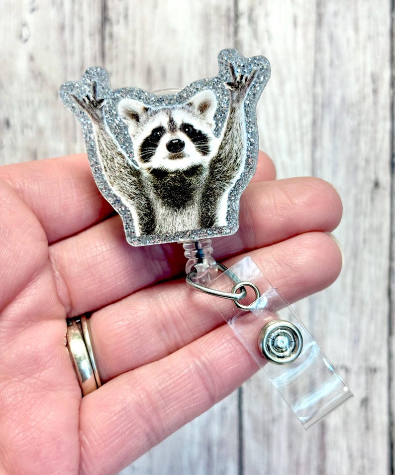 May include: A silver glitter badge reel with a raccoon wearing a surprised expression and raising its paws in the air. The raccoon is printed on a clear plastic circle.