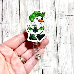 May include: A white duck wearing a green hat and green boots with shamrocks on its body. The duck is attached to a clear plastic retractable badge holder.
