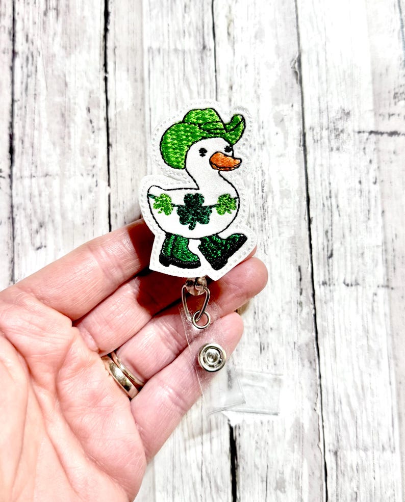 May include: A white duck wearing a green hat and green boots with shamrocks on its body. The duck is attached to a clear plastic retractable badge holder.