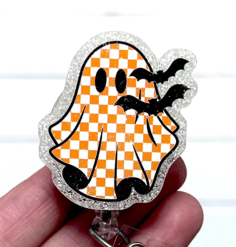 May include: A Halloween-themed badge reel featuring a smiling ghost with an orange and white checkered pattern. Two black bats are flying near the ghost. The badge reel has a glittery, clear border and a retractable cord.