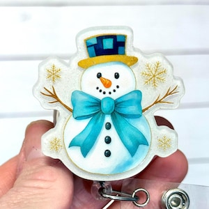 May include: A snowman-themed badge reel with a blue bow tie, black buttons, and an orange carrot nose. The snowman wears a yellow hat with a blue square pattern. The badge reel has a clear, glittery border with snowflake accents and brown twig arms.