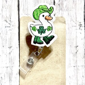 May include: A white duck wearing a green hat and green boots with shamrock designs. The duck is attached to a clear plastic badge holder.