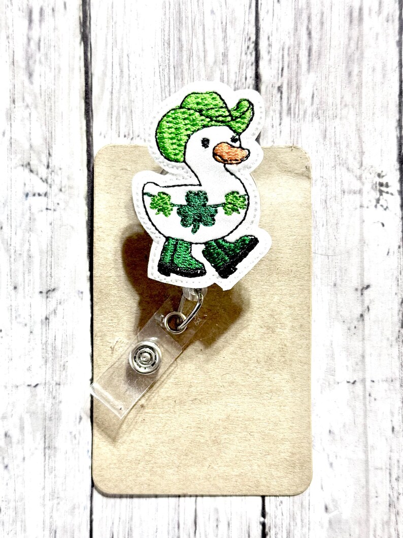 May include: A white duck wearing a green hat and green boots with shamrock designs. The duck is attached to a clear plastic badge holder.