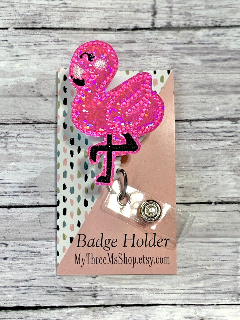 May include: Pink glitter flamingo badge holder with a clear plastic badge clip. The holder has a pink background with black and white polka dots. The text "Badge Holder" and "MyThreeMsShop.etsy.com" is printed on the holder.