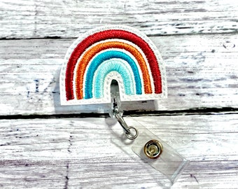 May include: A white rainbow-shaped badge holder with a clear plastic strap and a metal clip. The rainbow is embroidered with red, orange, yellow, blue, and green colors.