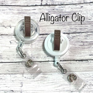 May include: Two white alligator clips with clear retractable badge holders. The clips have a silver metal finish and are attached to clear plastic straps.