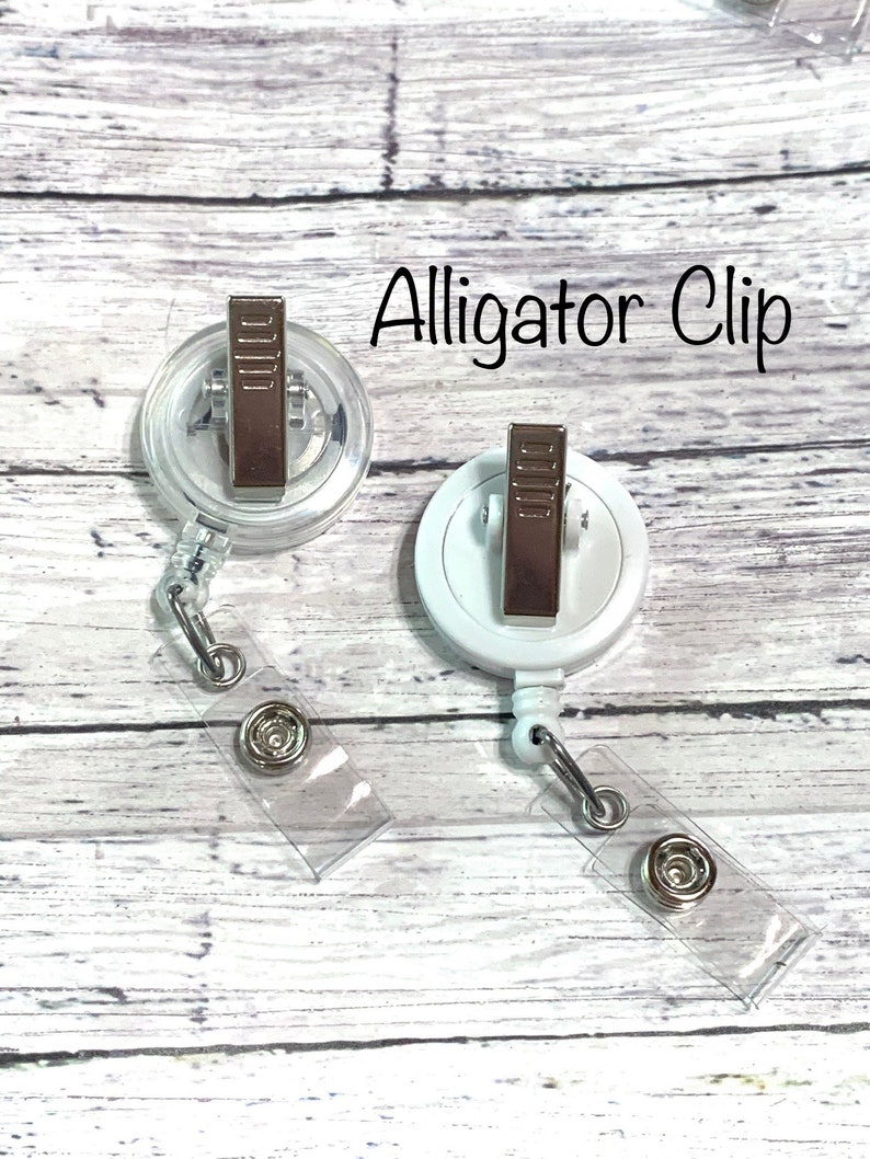 May include: Two white alligator clips with clear retractable badge holders. The clips have a silver metal finish and are attached to clear plastic straps.