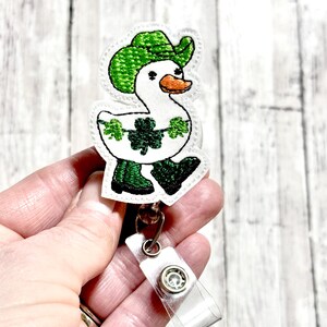 May include: White embroidered duck wearing a green hat and green boots with shamrocks. The duck is attached to a clear plastic badge holder with a silver clip.
