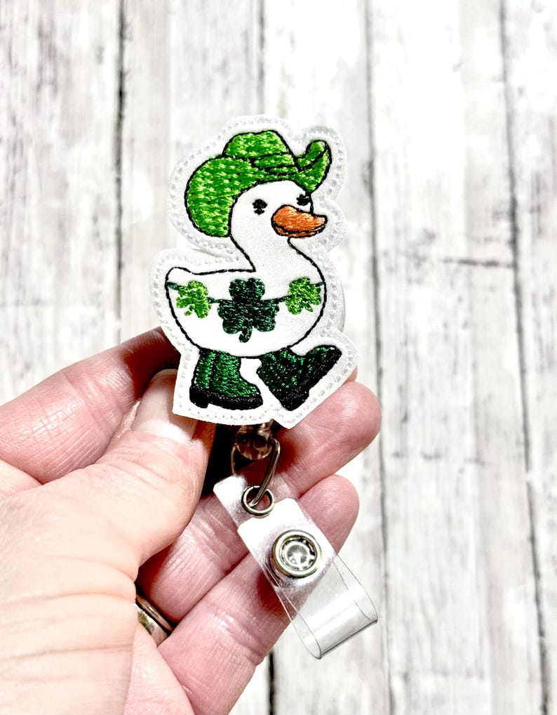 May include: White embroidered duck wearing a green hat and green boots with shamrocks. The duck is attached to a clear plastic badge holder with a silver clip.