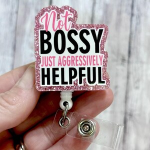 May include: A pink glitter badge reel with the words "Not Bossy Just Aggressively Helpful" in black and white. The badge reel has a white retractable cord and a clear plastic clip.