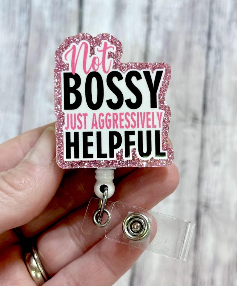 May include: A pink glitter badge reel with the words "Not Bossy Just Aggressively Helpful" in black and white. The badge reel has a white retractable cord and a clear plastic clip.
