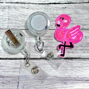 May include: Two clear retractable badge holders with a pink glitter flamingo charm. The badge holders have a clear plastic clip and a silver metal clip.