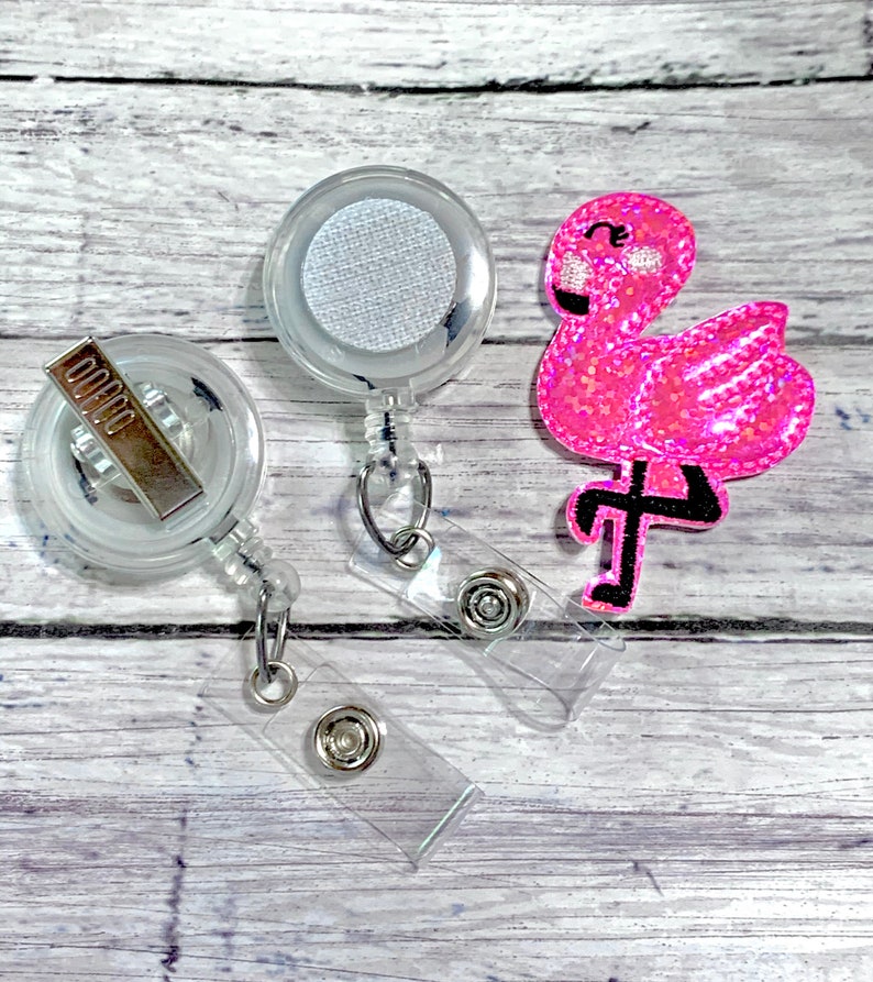 May include: Two clear retractable badge holders with a pink glitter flamingo charm. The badge holders have a clear plastic clip and a silver metal clip.
