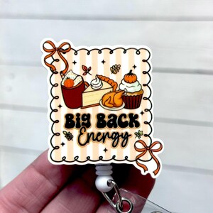 May include: A square badge reel with a Thanksgiving theme. The design features a cartoon illustration of a cup of coffee, pie, turkey, and a cupcake. The text "Big Back Energy" is in the center. The badge reel is white with a retractable cord.