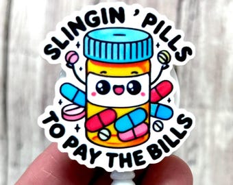 May include: A white badge reel with a cartoon pill bottle design. The bottle is yellow with a blue lid and has a smiling face. The text reads "SLINGIN' PILLS TO PAY THE BILLS". The design includes colorful pills.