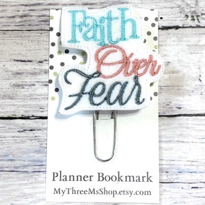 May include: A white planner bookmark with a blue and pink embroidered design that says "Faith Over Fear". The bookmark is attached to a silver binder clip.