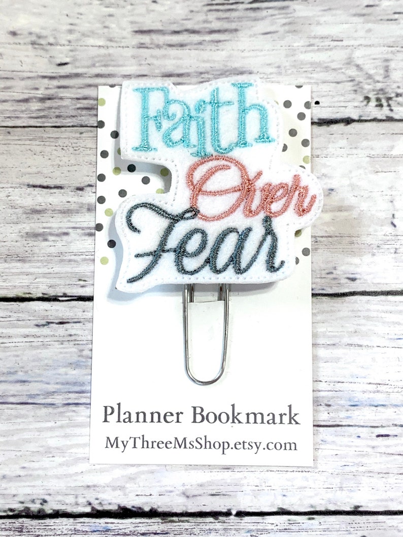 May include: A white planner bookmark with a blue and pink embroidered design that says "Faith Over Fear". The bookmark is attached to a silver binder clip.