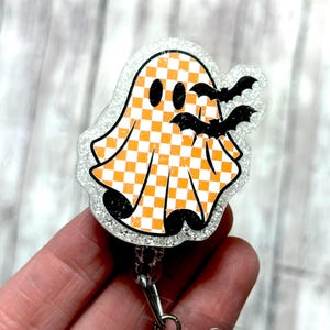 May include: A Halloween-themed badge reel featuring a ghost with an orange and white checkered pattern and black bats. The badge reel has a glittery border and a retractable cord.