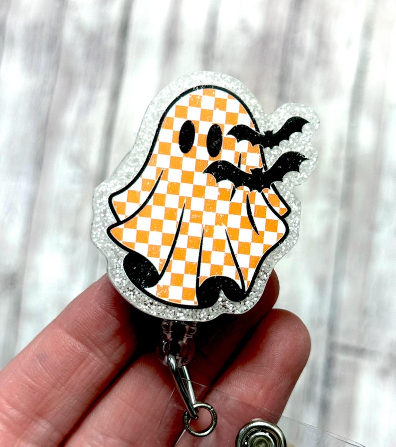 May include: A Halloween-themed badge reel featuring a ghost with an orange and white checkered pattern and black bats. The badge reel has a glittery border and a retractable cord.