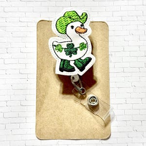 May include: A white goose wearing a green cowboy hat and green boots with shamrocks around its neck. The goose is attached to a retractable badge holder.